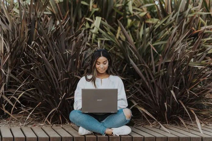 Woman sitting on planks using a laptop in front of tall grass